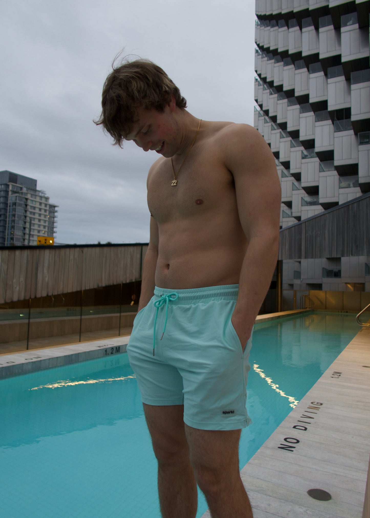 Man in light blue luxury matching swim shorts standing by a pool with a Vancouver cityscape in the background