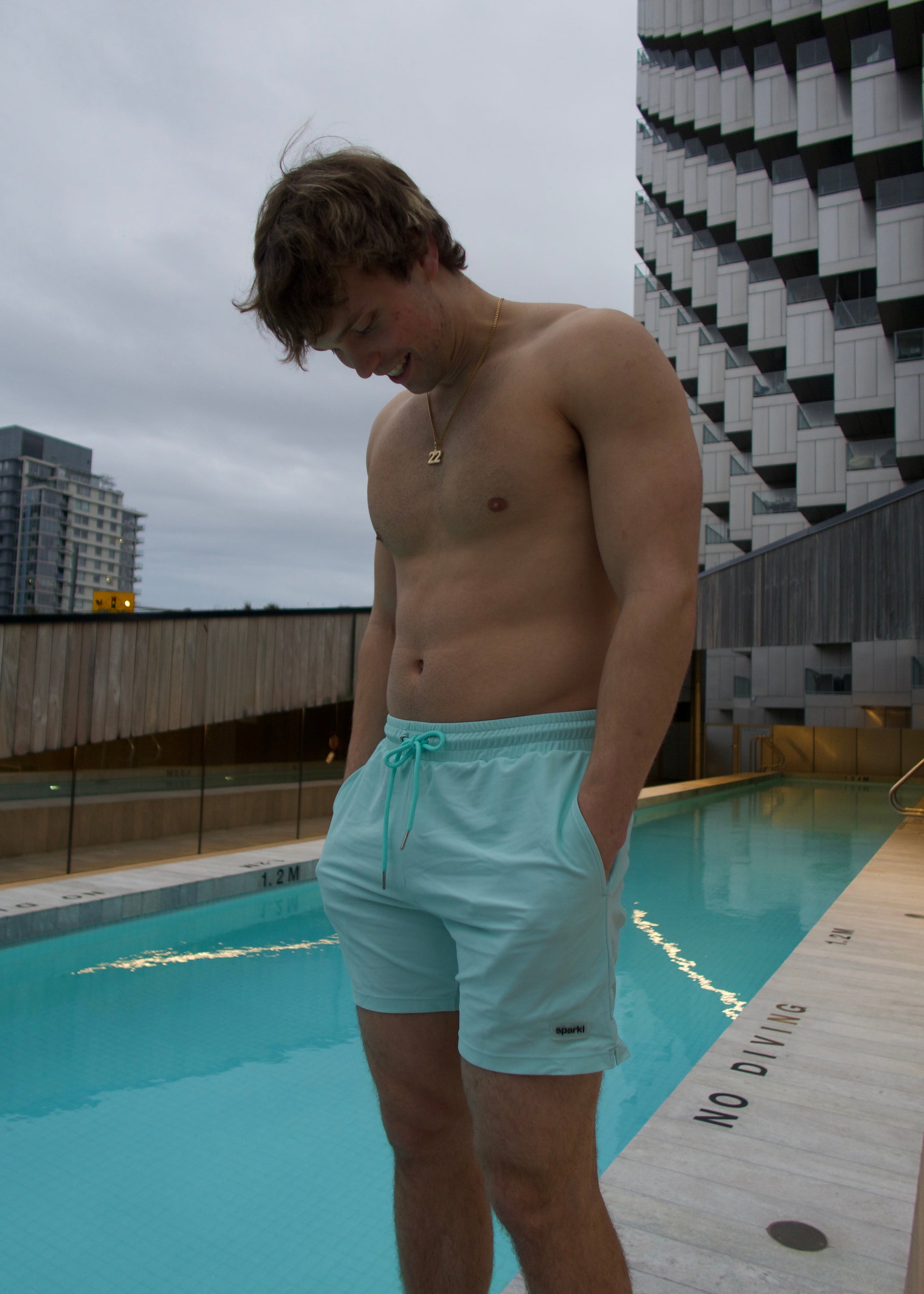 Man in light blue luxury matching swim shorts standing by a pool with a Vancouver cityscape in the background