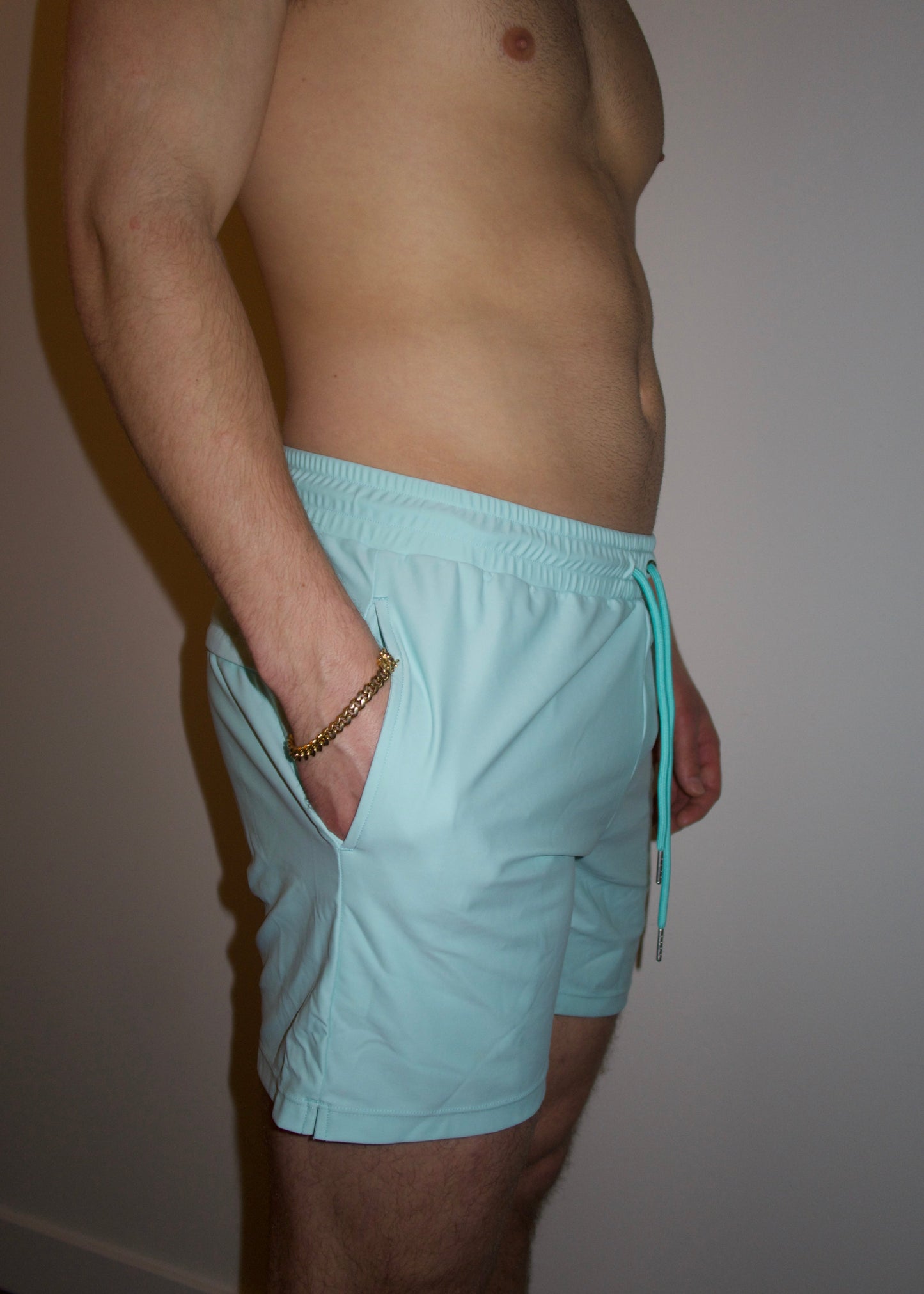 Male with hand in pocket wearing light blue swim shorts against white wall. 